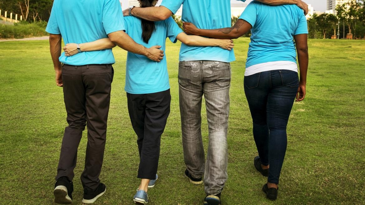 Seen from behind, four people walk across an outdoor grass field with their arms across each others' backs and shoulders.